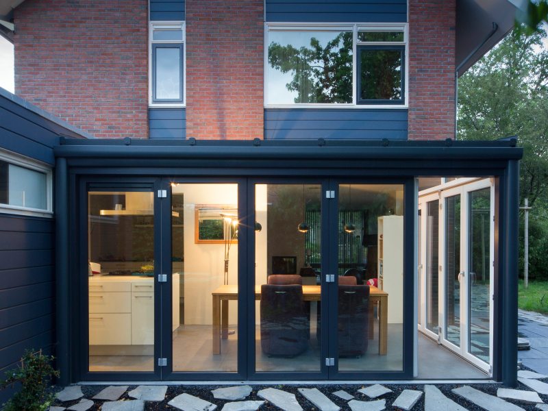 Kwaliteit kunststof Een buitenfoto van de aanbouw van een modern huis met grote, glazen vouwdeuren en een stenen pad in de achtertuin.