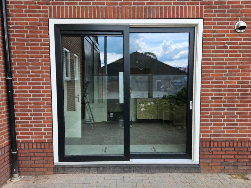 Kwaliteit kunststof Een kunststof schuifpui met een zwart frame en een wit buitenkozijn, geplaatst in een rode bakstenen muur. De pui weerspiegelt de lucht en de overkant van de tuin.